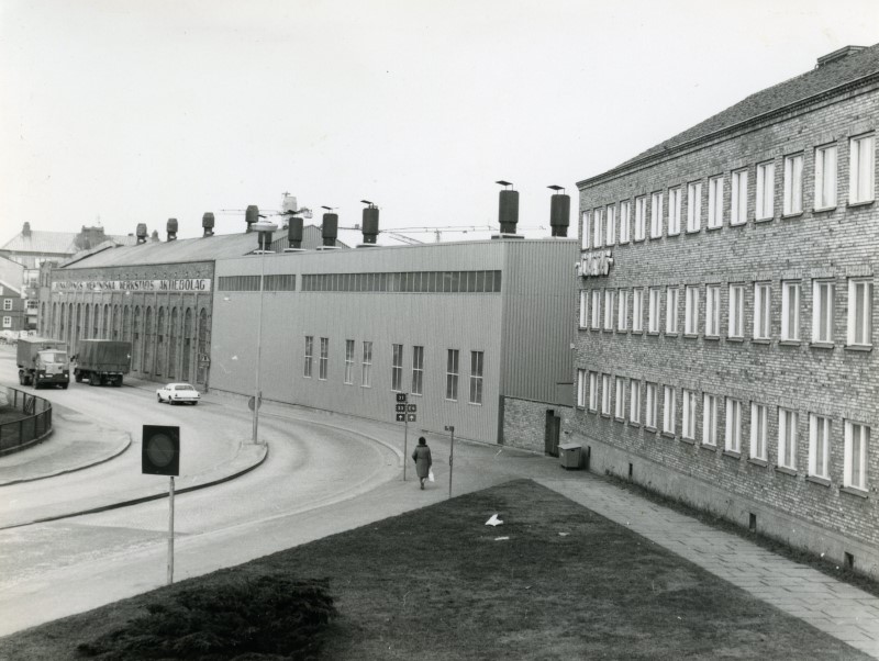 Monochrome photograph of the foundry hall and the extension as it was in 1969. In the street next to the building two heavy trucks can be seen and a small car.