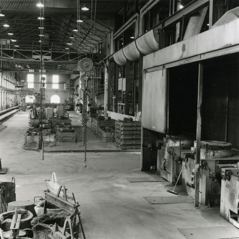 Monochrome photograph of the foundry hall interior some time between ~1975 and ~1990. In the photo machines and materials can be seen on the factory floor.