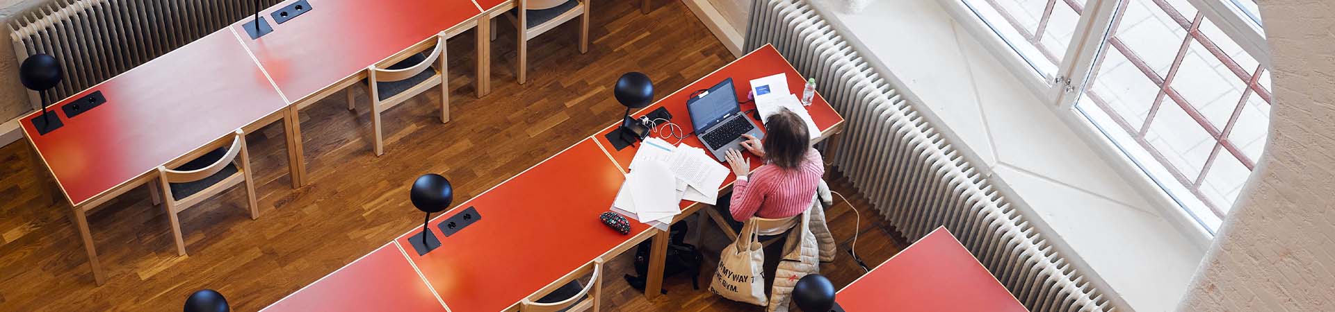 A lone student studying with a laptop and lots of documents spread out on a red table. The table is next to a large window. The picture is taken from above.