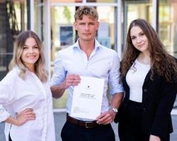 Fanny Johansson, Linus Högbäck and Austra Kase stand together with JIBS' entrance behind them. Linus is in the centre and holds their winning thesis in his hands. 