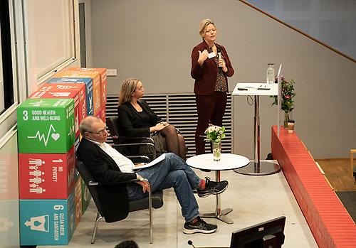 Amalie Kestler stands on stage in a lecture room. She has a microphone in her hand that she is talking into. Seated to the left on two chairs are Lotta Edling and Stefan Melesko. 