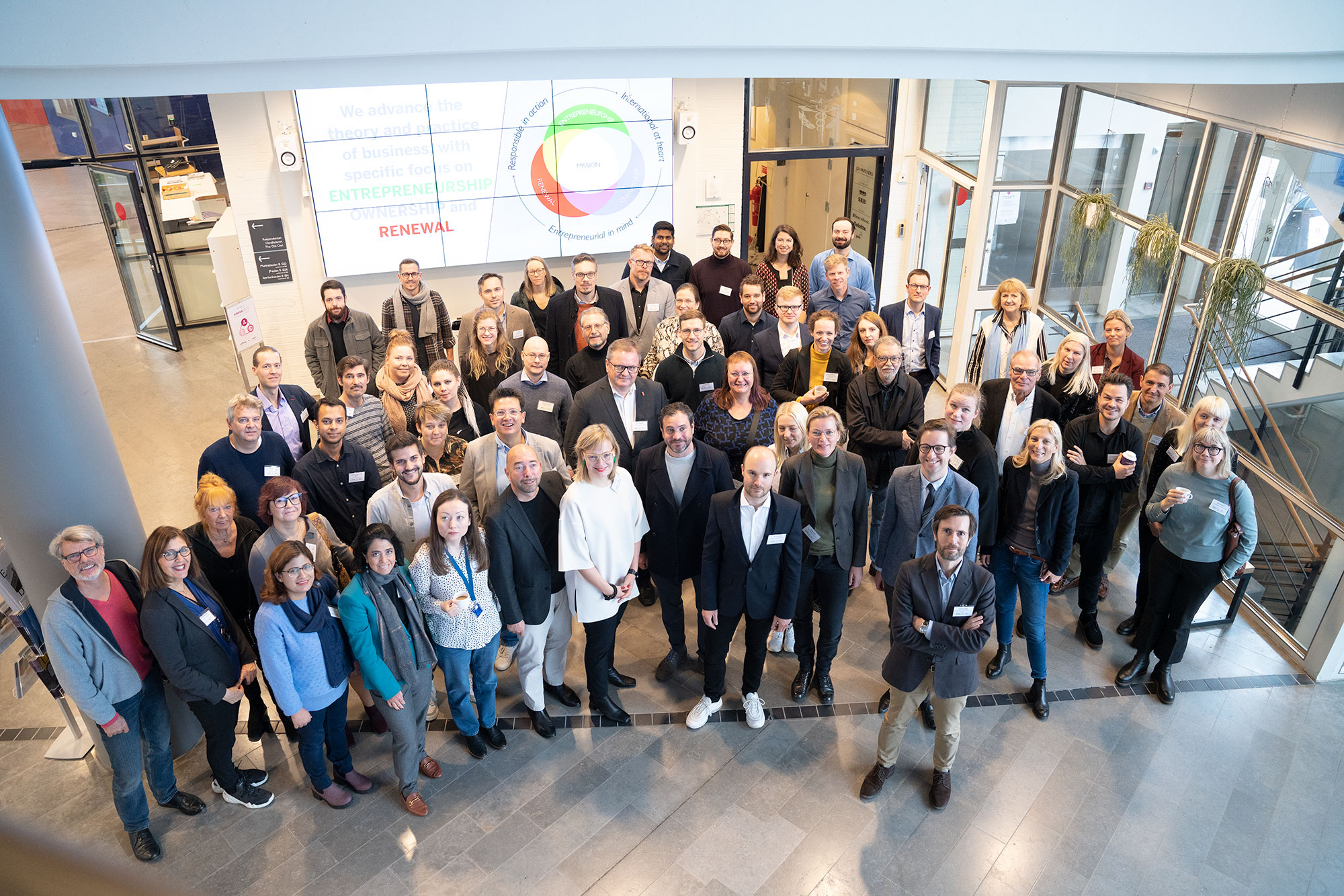 A group of aproximately 90 people stand in the entrance of a lobby at a business school. They are looking up towards the camera. 