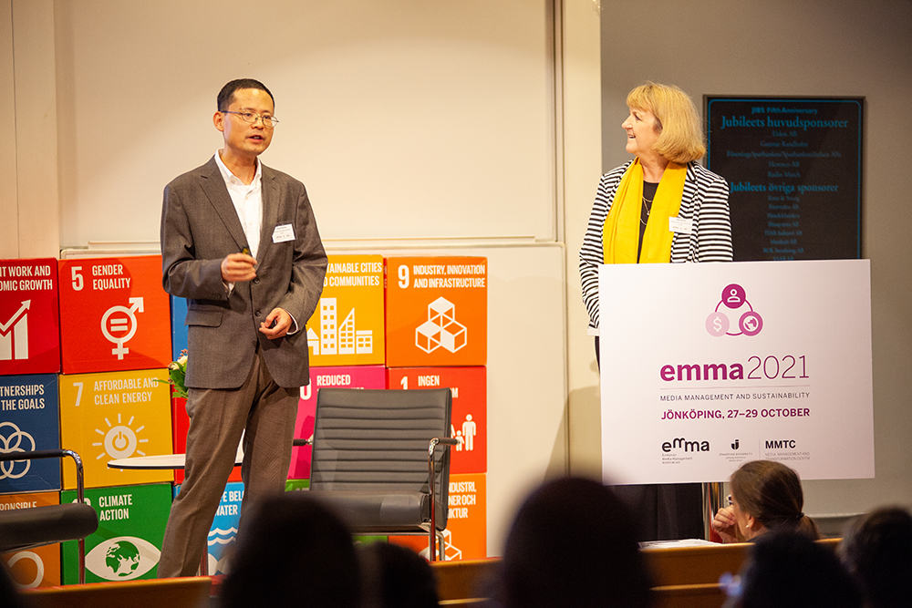 A male and female researcher stand on stage. One is talking, the other is turned to look at him. In the foreground is an audience sitting in lecture hall fold down seats. 