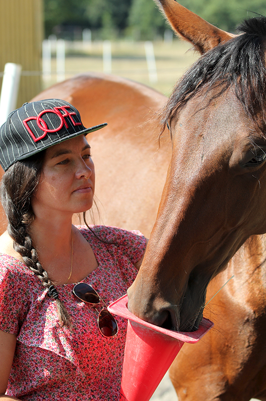 En kvinna med keps och långt mörkt hår i en fläta håller en plastkon och en brun häst nosar i. 