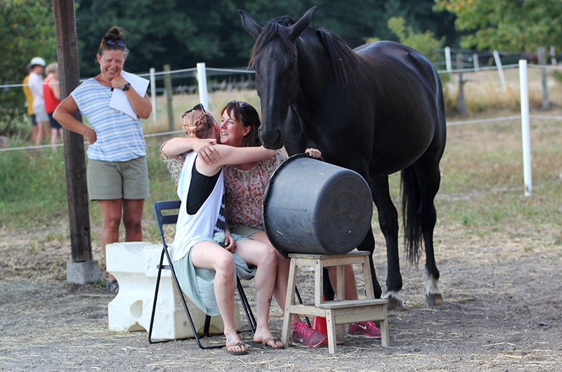 Två kvinnor sitter på stolar i en hästhage och kramar om varandra. En mörk häst står och vilar intill. I bakgrunden syns flera människor. 