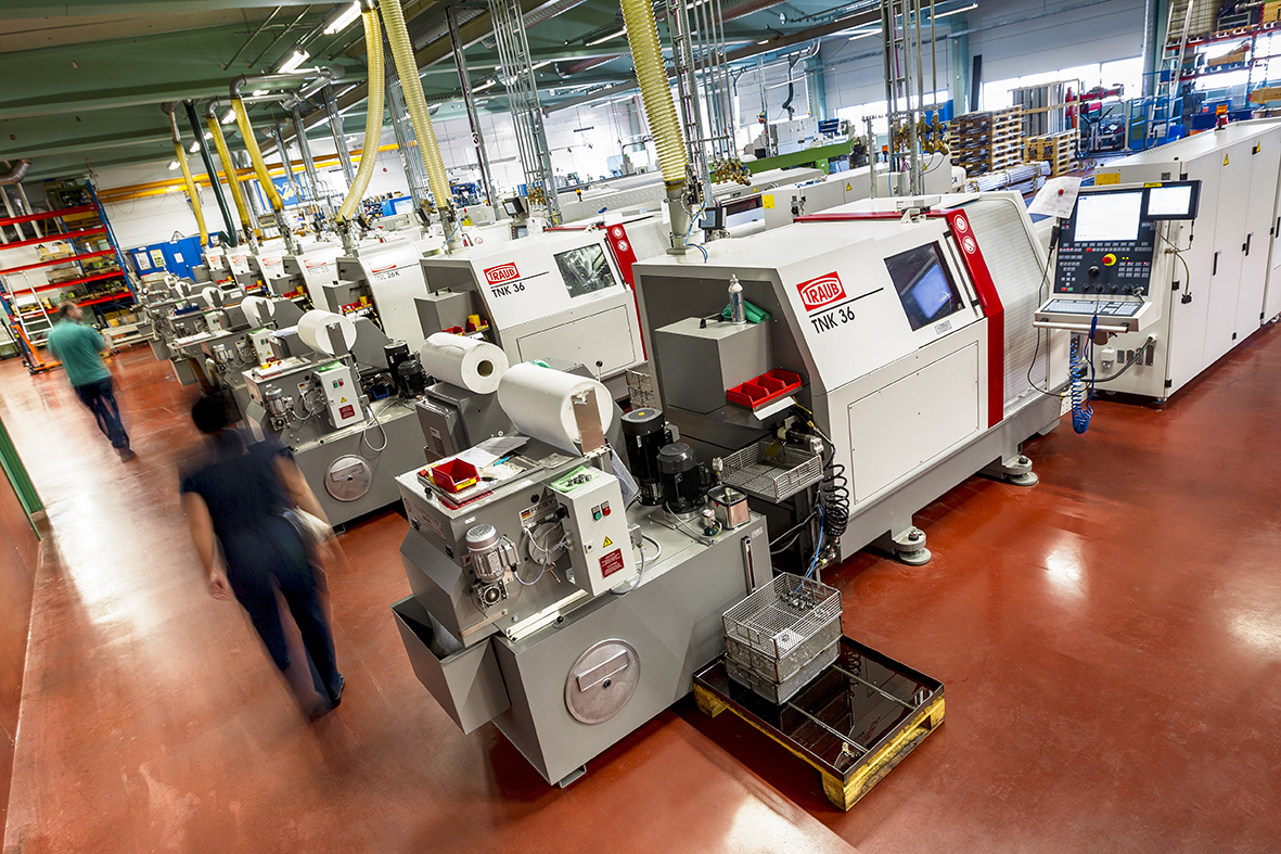 Image shows the inside of the factory floor at Gnosjö Automatsvarvning. There are a series of metal turning machines and two members of staff to the left of the image. 