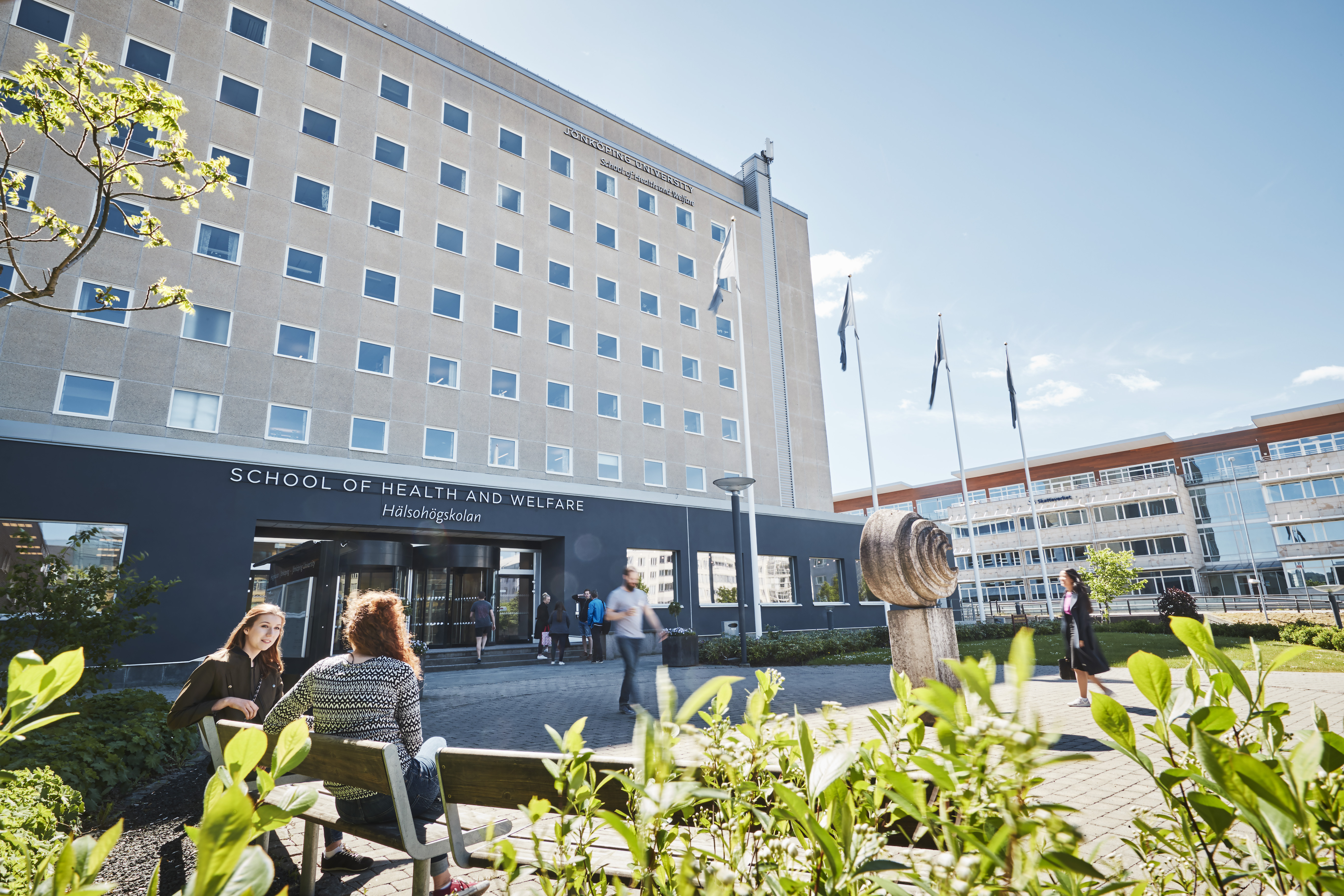 Hälsohögskolans framsida med grönskande blad och två personer som sitter på en parkbänk i förgrunden