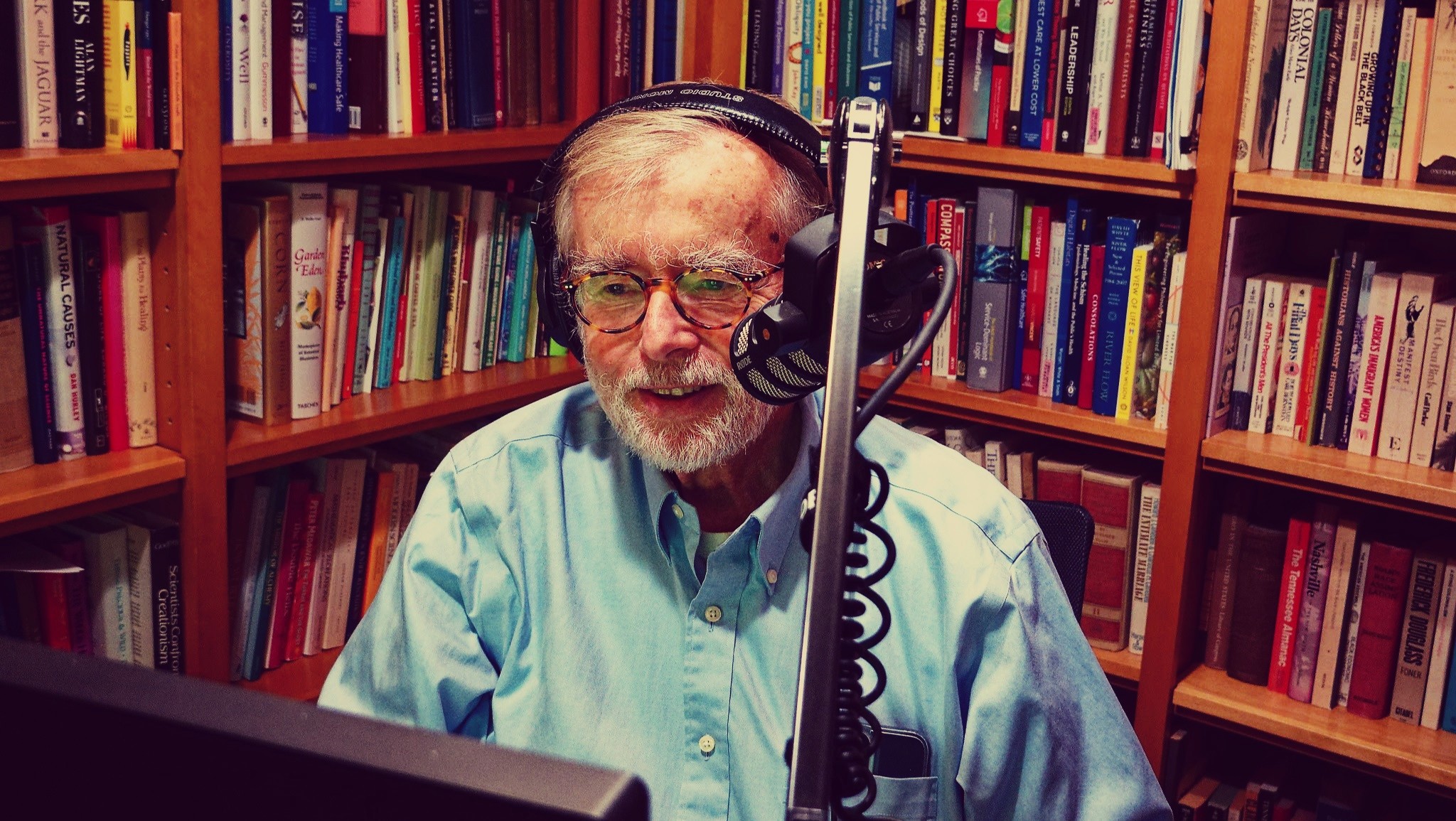 Paul Batalden smiling and sitting in front of a computer in his library, wearing headphones and speaking into a microphone