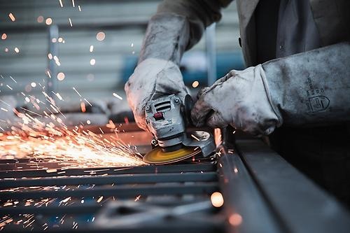 närbild av armar och händer på en metallarbetare i en fabrik, som håller ett metallslipverktyg med gnistor som kommer från den.