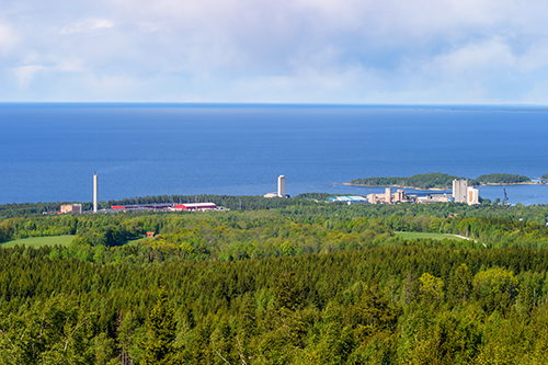 utsikt över svensk skog med havet i bakgrunden. På medelavstånd nära kusten ligger en fabrik.